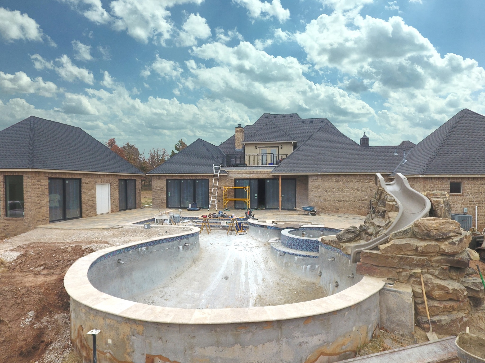 Large estate pool under construction with travertine coping, rock slide feature, and brick home backdrop