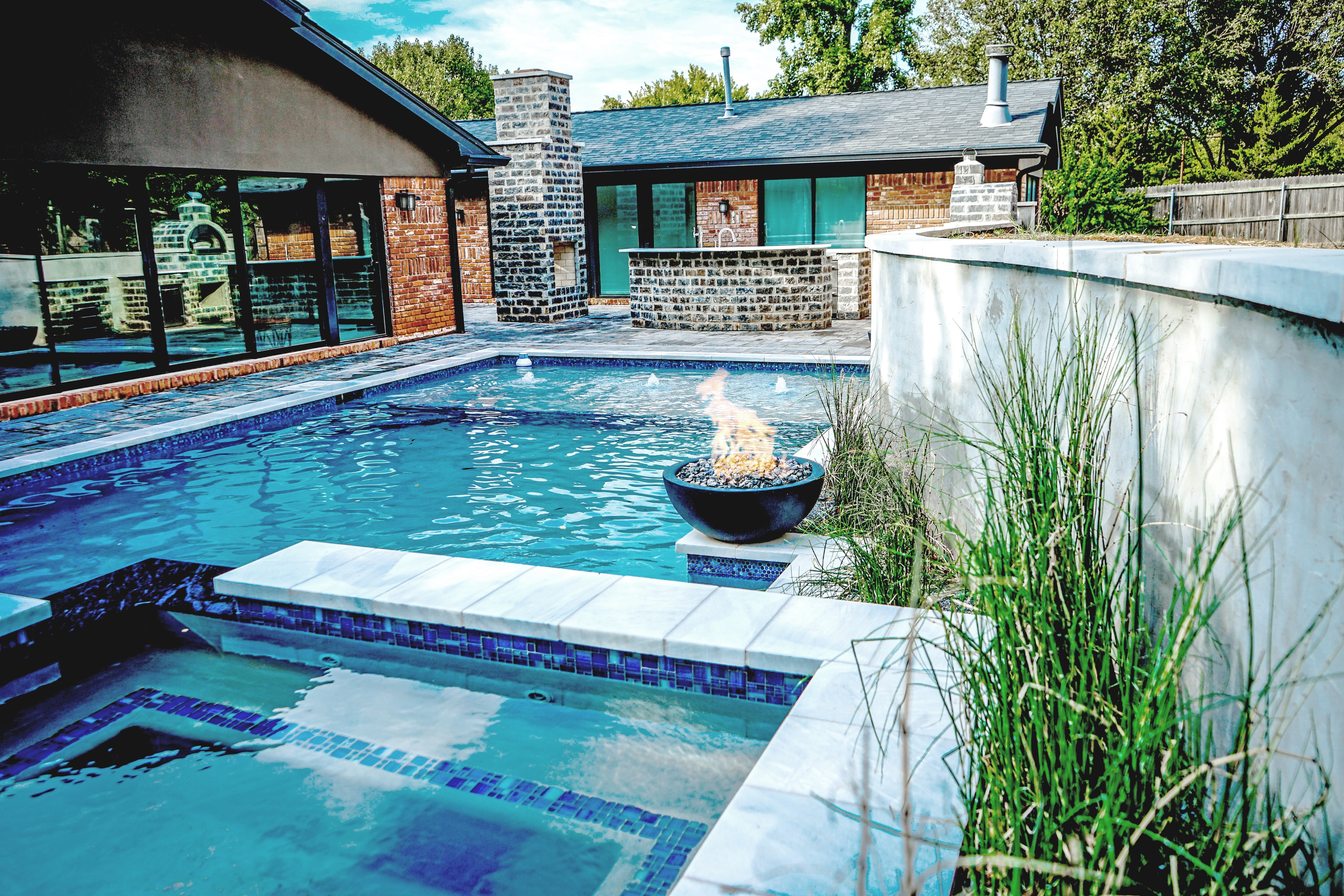 Modern rectangular pool with in-pool fire bowl, raised spa, and brick outdoor kitchen beyond