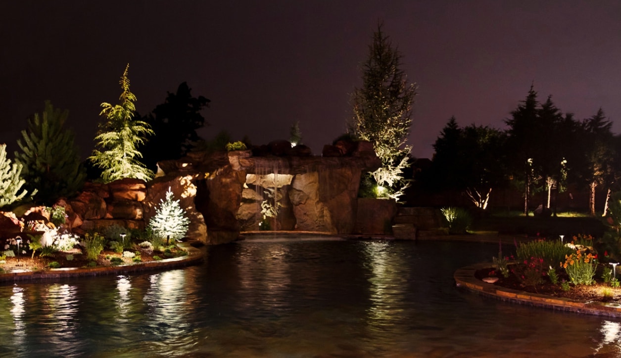 Illuminated grotto pool with waterfall, rock formations, and landscape lighting at night