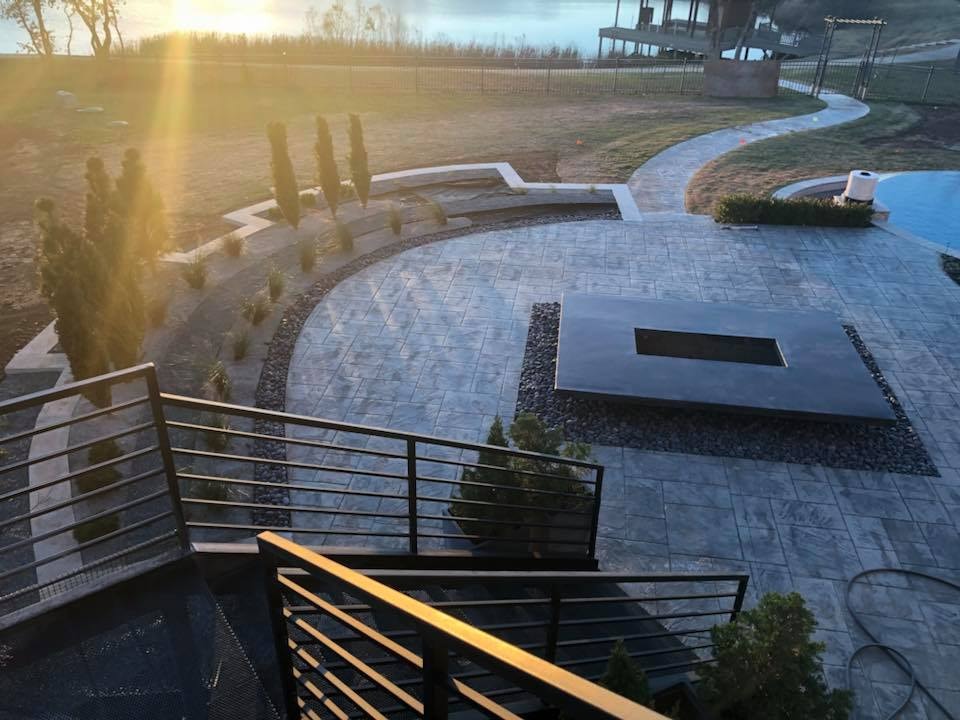 Stamped concrete lakefront patio with recessed fire table, Italian cypress plantings, and golden sunset over the water