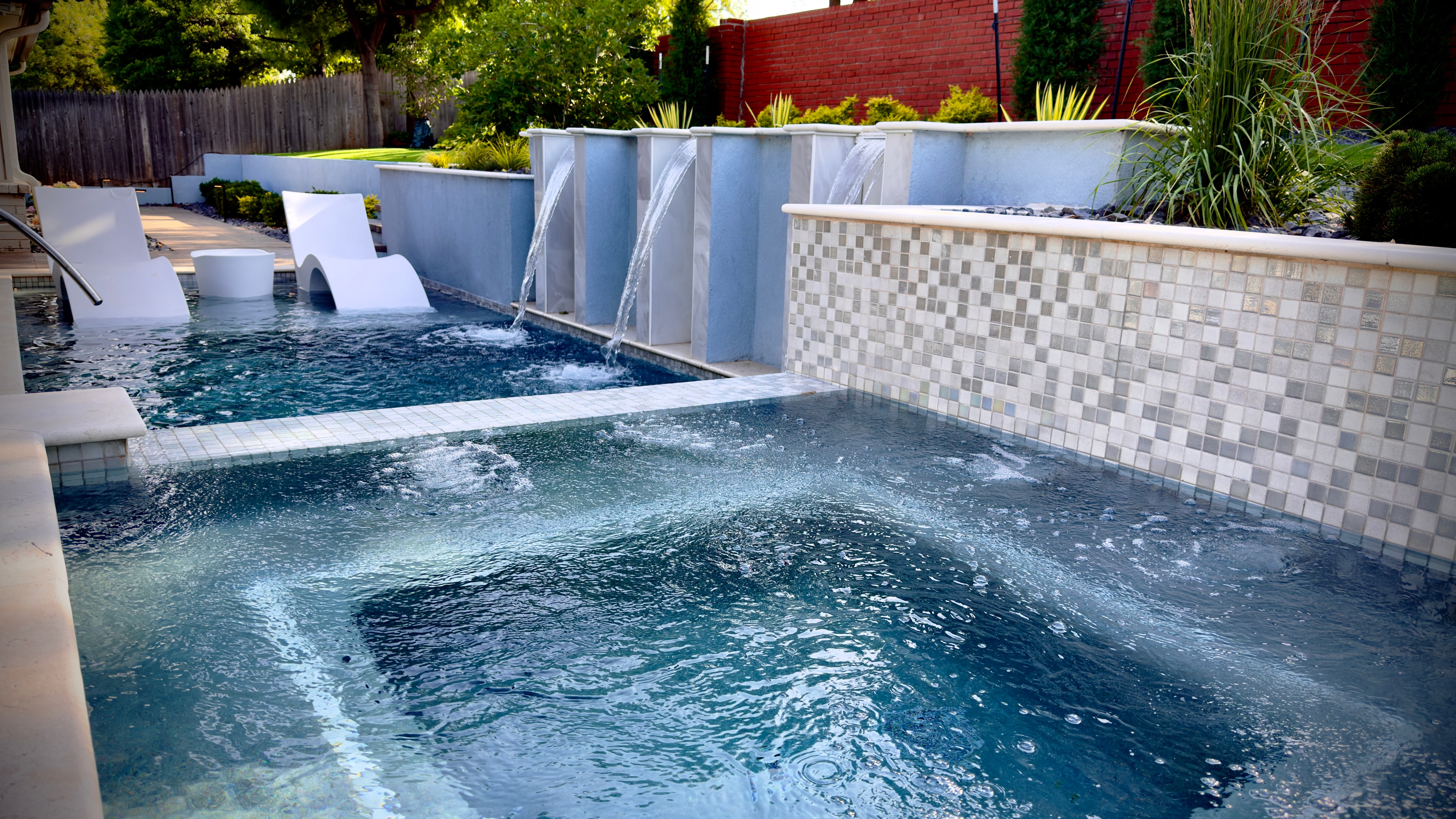 Modern pool with mosaic tile waterfall wall, white chaise lounges, and lush plantings