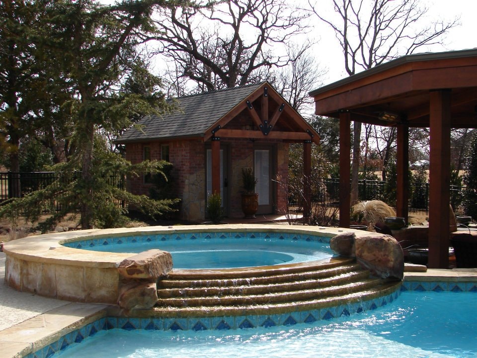 Raised spa with cascading steps into pool beside brick poolhouse and cedar pergola