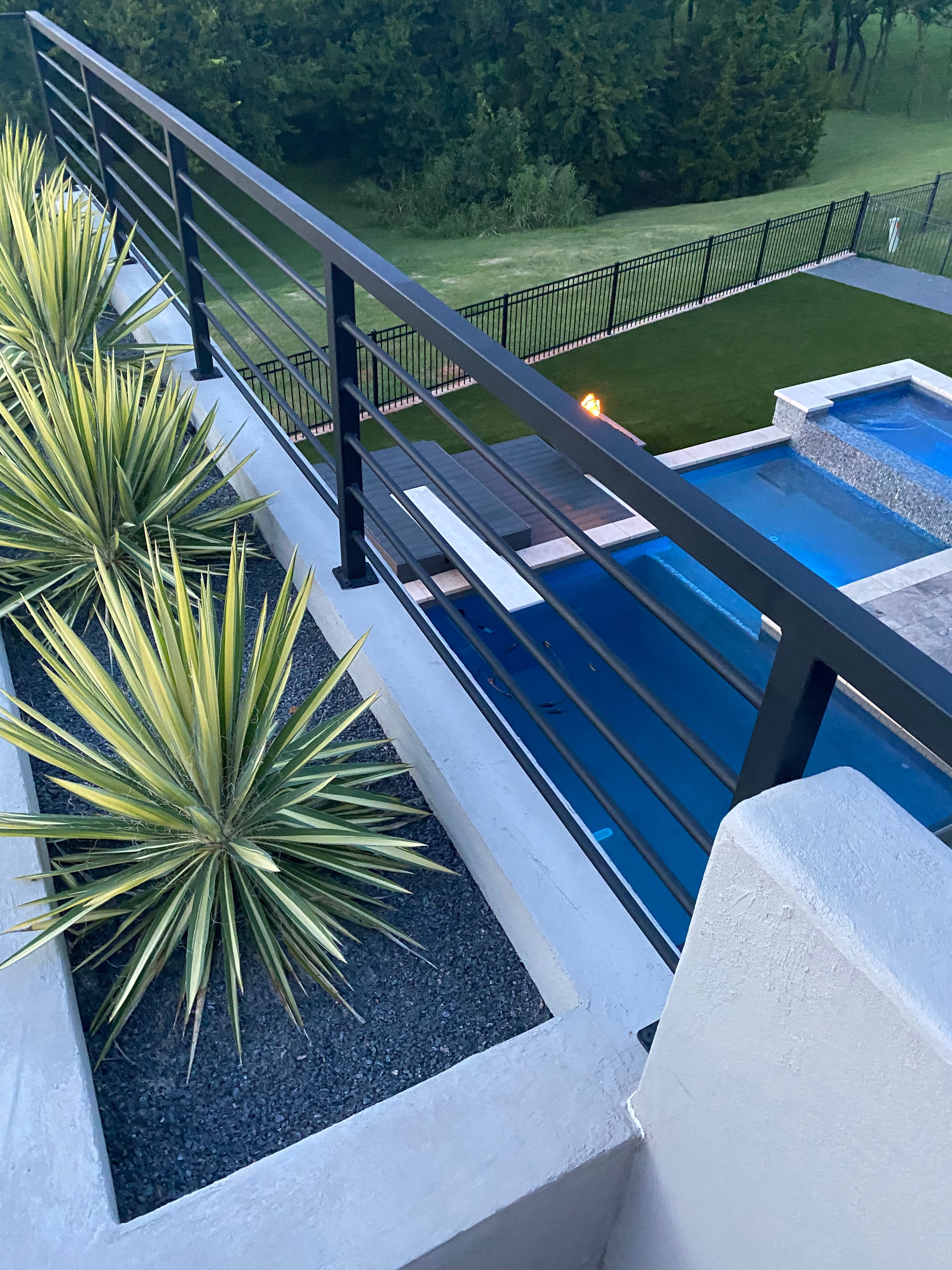 Elevated lap pool with steel horizontal railing, yucca planting bed, and putting green below at dusk