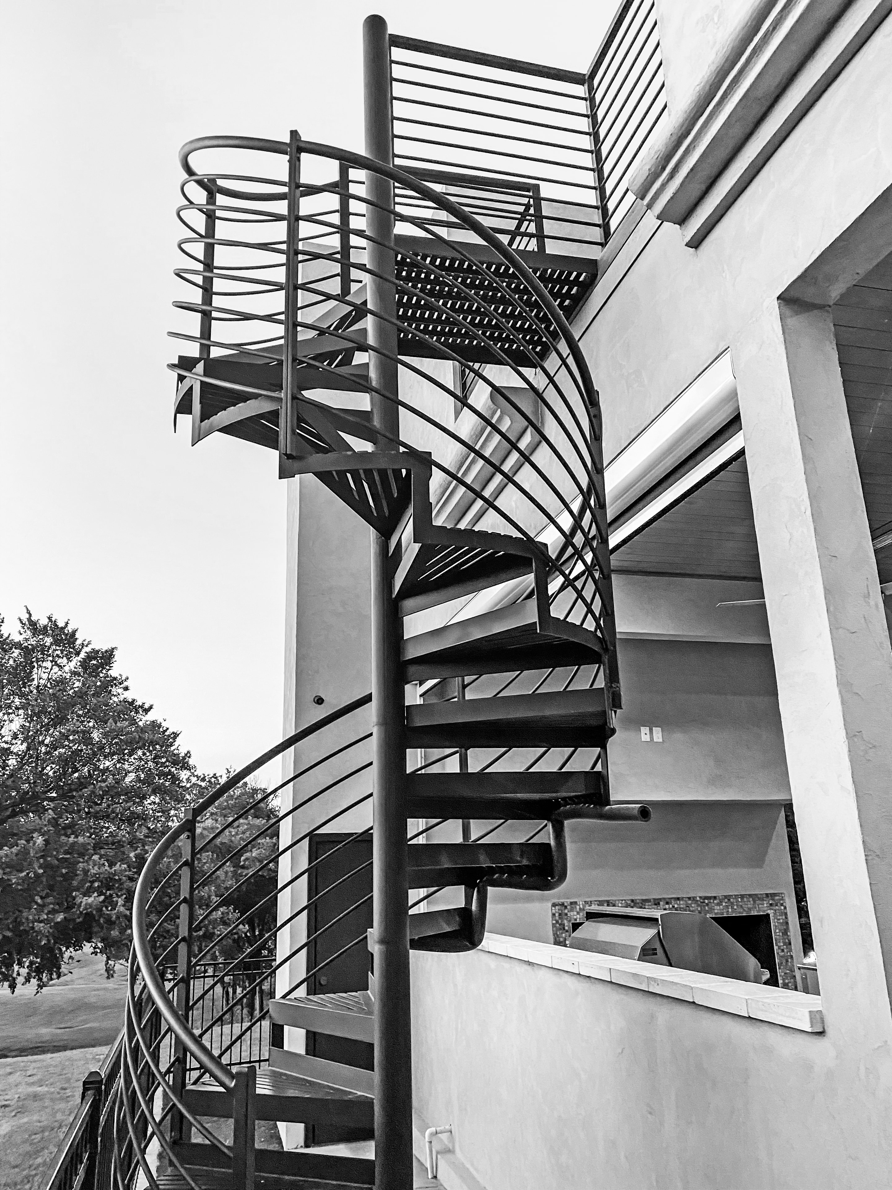 Black-and-white photograph of dramatic exterior spiral staircase with steel railings and outdoor kitchen below