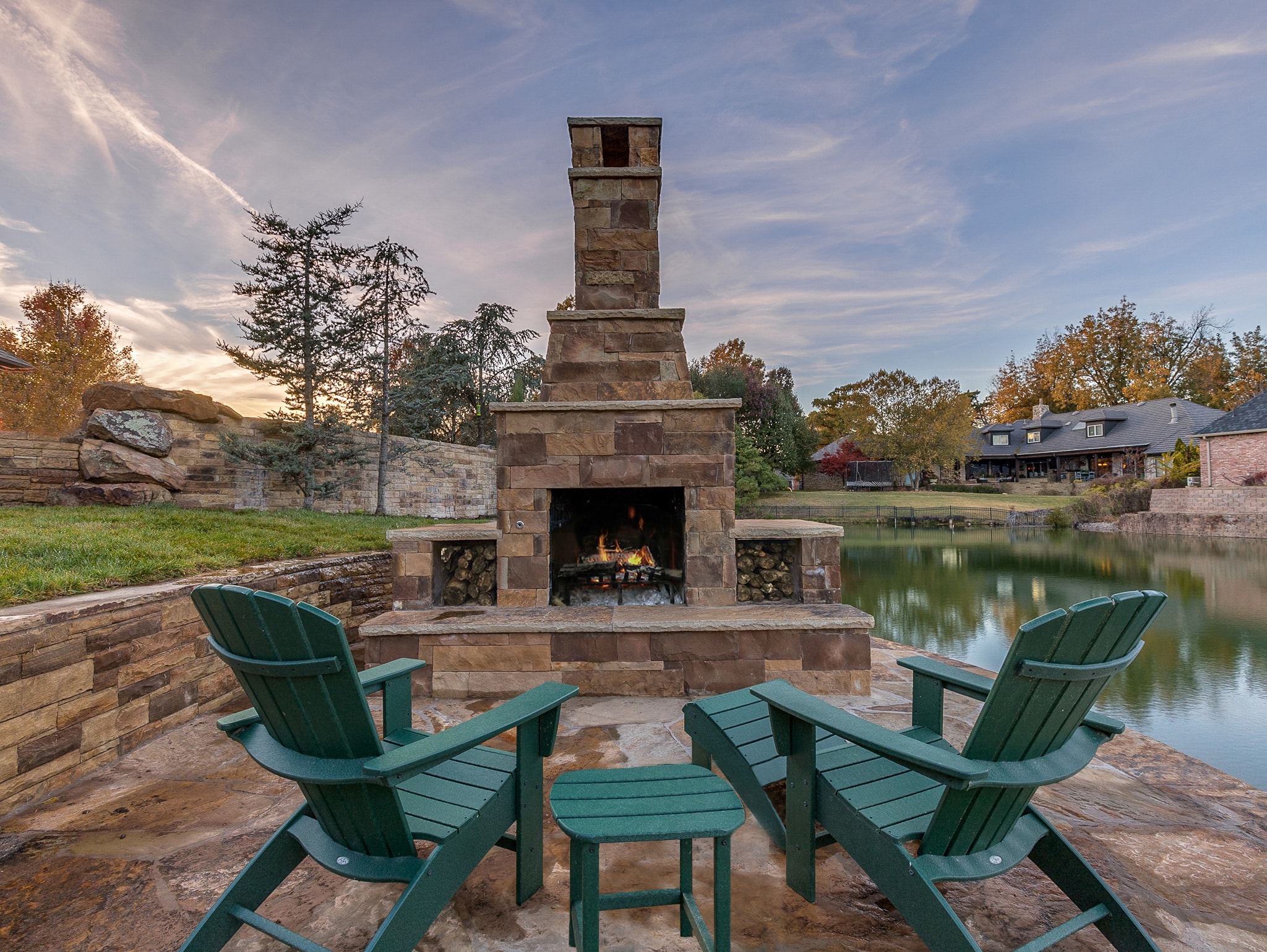 Stone outdoor fireplace with Adirondack chairs on flagstone patio beside private pond at sunset