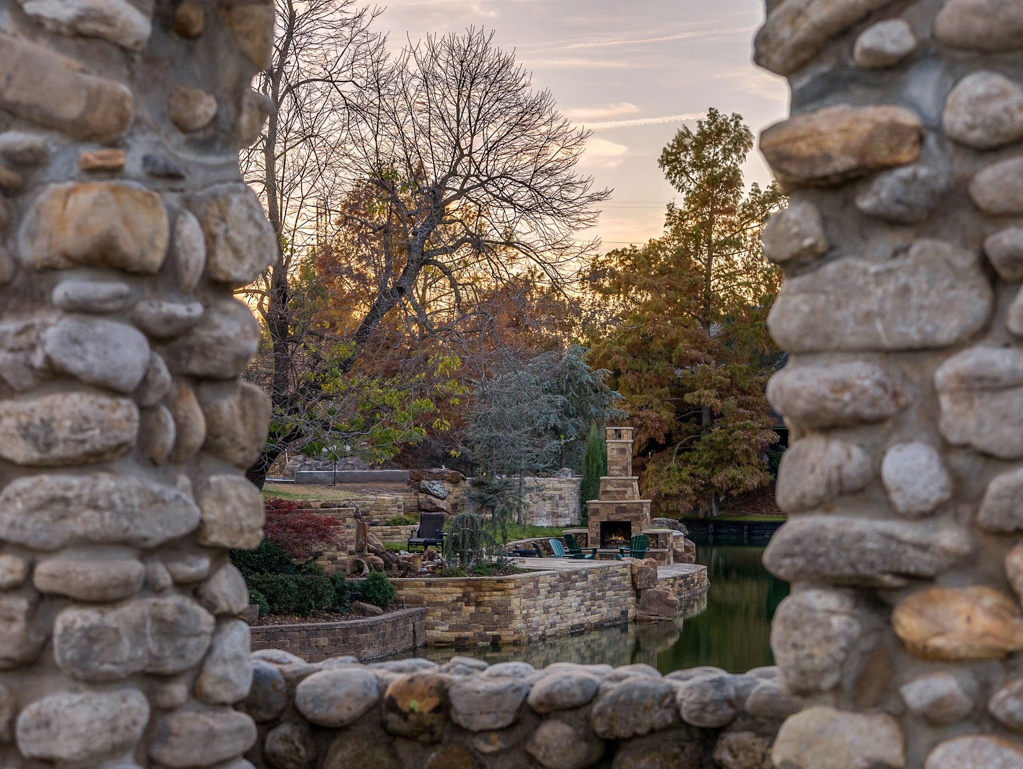 Framed view through stone arch of terraced retaining walls, fireplace, and pond at sunset